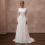 A woman stands in a studio wearing a long-sleeve white wedding gown with an embellished lace bodice and a flowing chiffon skirt, posed against a textured beige wall and dark wood floor.