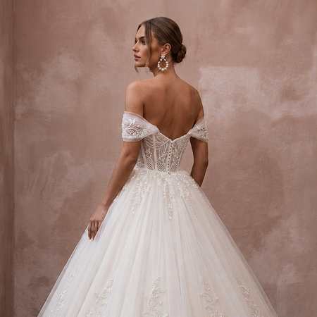 A woman wearing a voluminous off-the-shoulder wedding gown with lace embroidery and a full tulle skirt, photographed in a softly lit interior with a textured blush wall and wood floor.