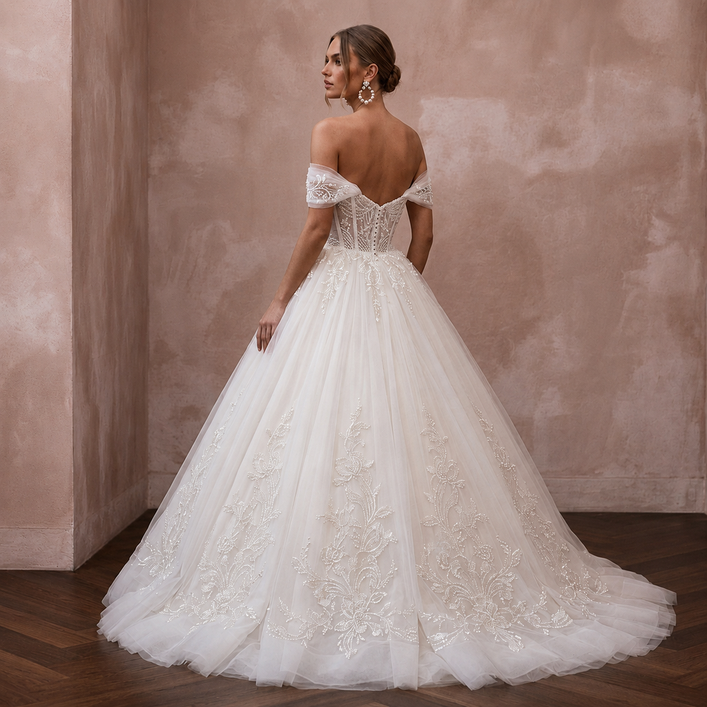 A woman wearing a voluminous off-the-shoulder wedding gown with lace embroidery and a full tulle skirt, photographed in a softly lit interior with a textured blush wall and wood floor.