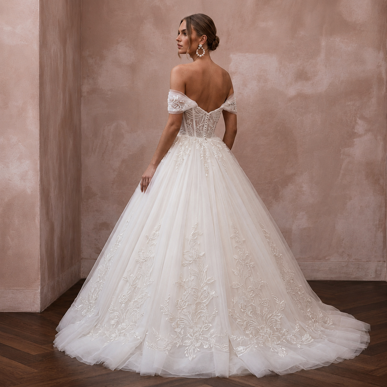 A woman wearing a voluminous off-the-shoulder wedding gown with lace embroidery and a full tulle skirt, photographed in a softly lit interior with a textured blush wall and wood floor.