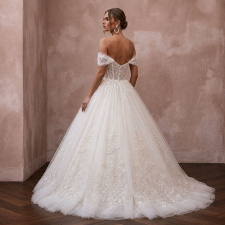 A woman wearing a voluminous off-the-shoulder wedding gown with lace embroidery and a full tulle skirt, photographed in a softly lit interior with a textured blush wall and wood floor.