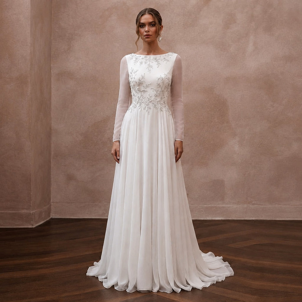 A woman stands in a studio wearing a long-sleeve white wedding gown with an embellished lace bodice and a flowing chiffon skirt, posed against a textured beige wall and dark wood floor.