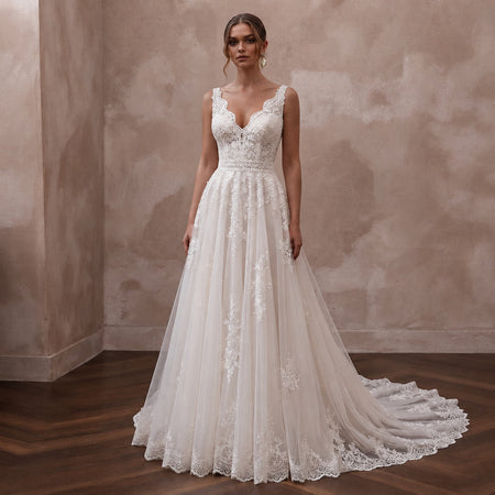 A woman modeling a sleeveless A-line wedding gown with a deep V-neckline, lace bodice, and a long sheer tulle train with floral lace applique, standing in a room with a textured beige wall and dark wood floor.