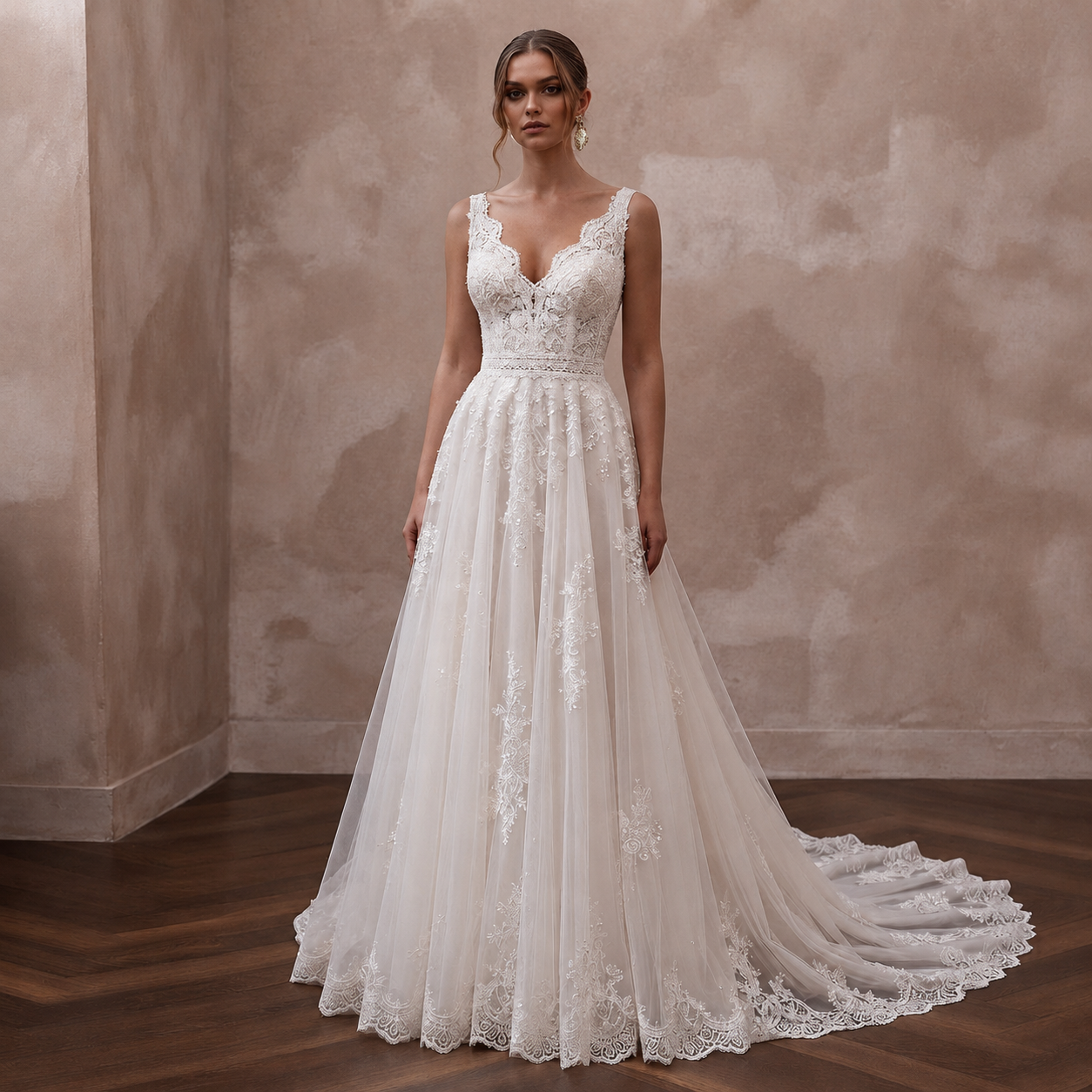 A woman modeling a sleeveless A-line wedding gown with a deep V-neckline, lace bodice, and a long sheer tulle train with floral lace applique, standing in a room with a textured beige wall and dark wood floor.