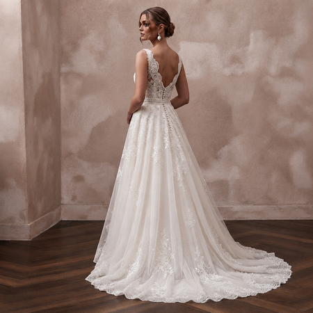 A woman wearing a white lace wedding gown stands with her back to the camera in an indoor setting with a textured beige wall and dark wood floor.