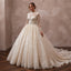 A woman modeling a voluminous ivory wedding gown with long lace sleeves, a high neckline, and a full tulle skirt with a train, posed in an indoor studio setting with a textured beige wall and dark wood floor.
