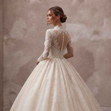 A woman wearing a voluminous ivory lace ball gown with long sleeves and a high neck, shown from the back in an indoor setting with a textured beige wall and dark wood floor.